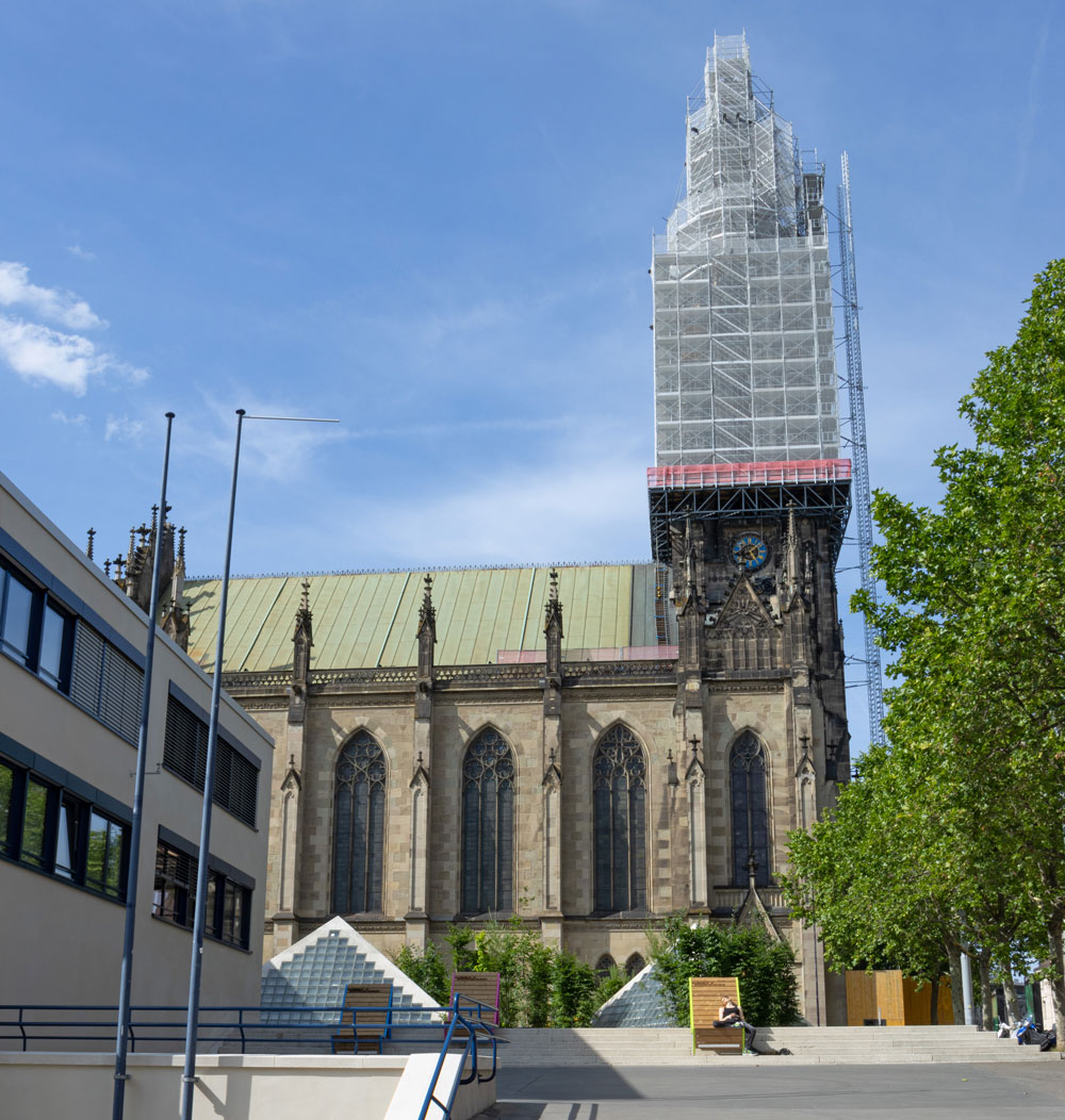 Seitliche Aufnahme der Elisabthenkirche mit dem Thaterplatz und den Pyramiden. Der Turm ist im Gerüst, vor der Kirche schläft ein Mann auf einer Bank.